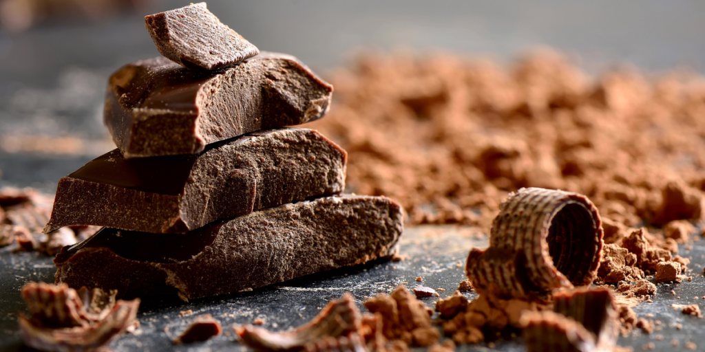 Des morceaux de chocolat noir empilés, entourés de copeaux de chocolat sur une surface sombre. Le chocolat est connu pour être une source riche en magnésium.