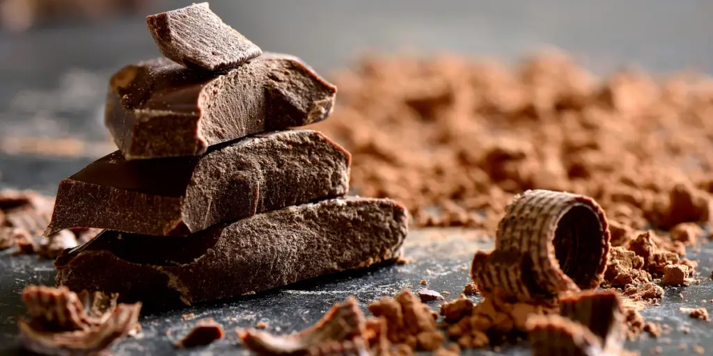 Des morceaux de chocolat noir empilés, entourés de copeaux de chocolat sur une surface sombre. Le chocolat est connu pour être une source riche en magnésium.